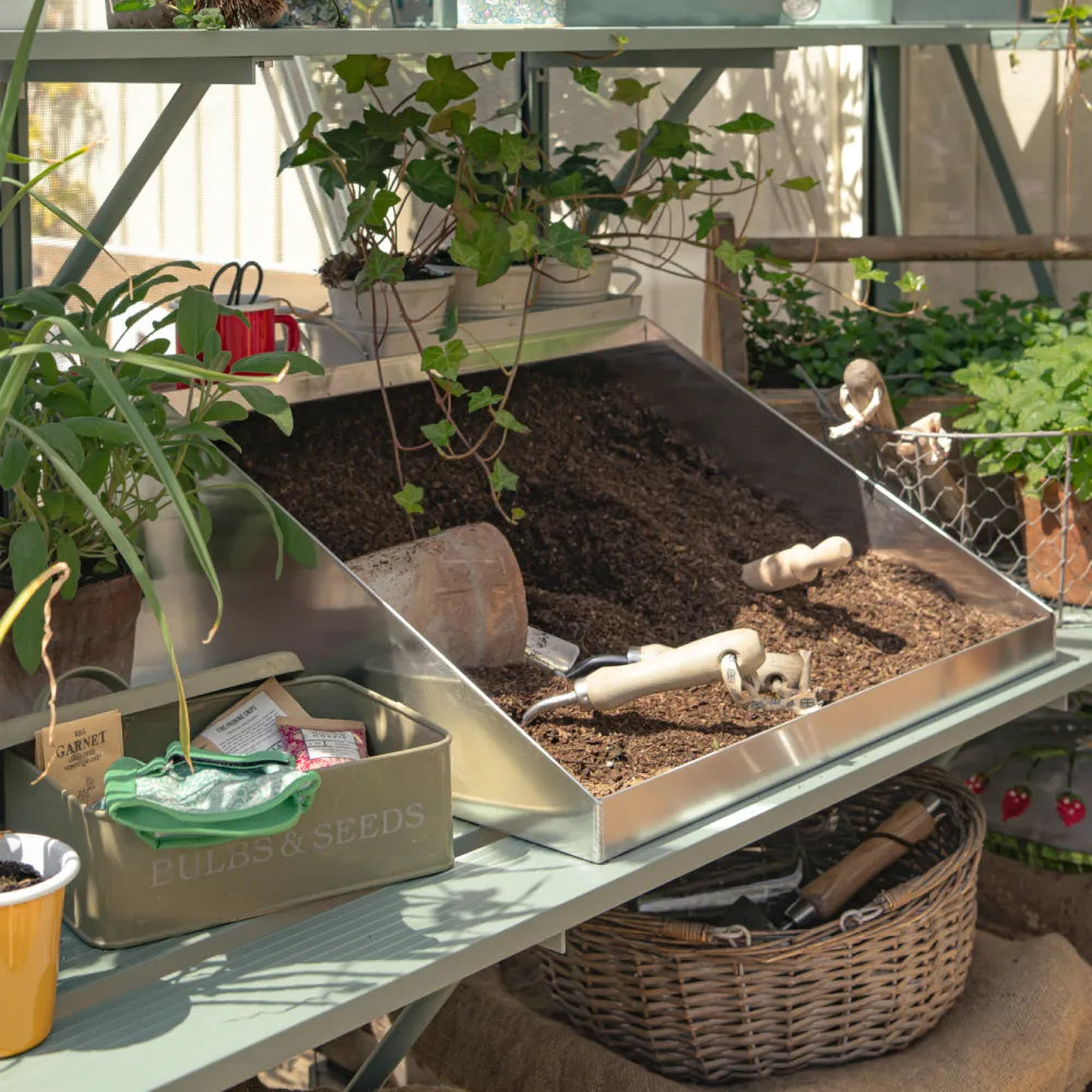 Soil sits in a metal planting tray alongside gardening tools, surrounded by plants and pots in a Rhino Greenhouse. A tin labeled BULBS & SEEDS holds seed packets.