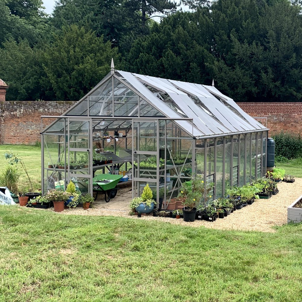 A glass Rhino Greenhouse shelters various plants on a gravel path, surrounded by more potted plants and a wheelbarrow, situated against a backdrop of trees and a brick wall.