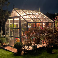 A glass Rhino Greenhouse illuminated at night, displaying various potted plants inside. It is surrounded by lush garden foliage and pots, creating a serene, well-maintained garden atmosphere.