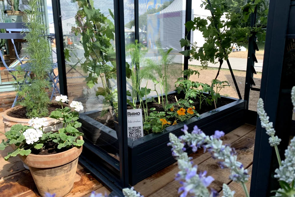 A glass Rhino Greenhouse holds various plants, including flowers and vegetables, displayed on a wooden deck. A sign reads Rhino Rhino Greenhouses with a QR code nearby.