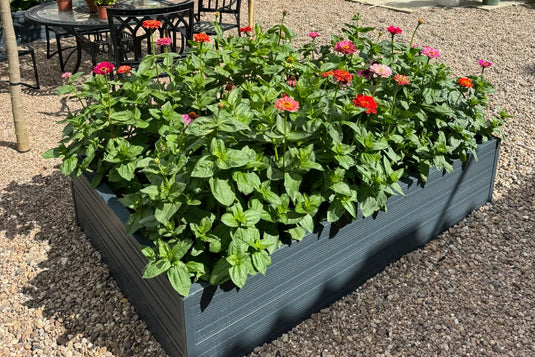 A rectangular, elevated planter box filled with vibrant pink, red, and orange flowers, situated on a gravel-covered outdoor patio beside a metal table and chairs.