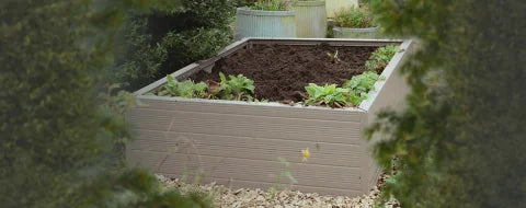 A rectangular Rhino metal raised garden bed filled with dark soil and small green plants is surrounded by lush foliage and additional metal planters in a garden setting.