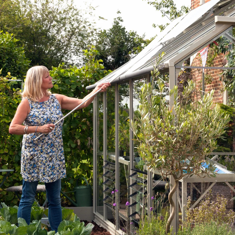 A woman stands by a Rhino Greenhouse, adjusting a roof vent with a rod. She is surrounded by lush greenery and various plants on a sunny day.