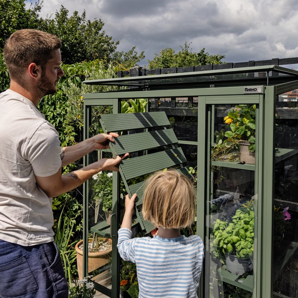 A man and a child install a louvered window on a Rhino Rhino Greenhouse surrounded by lush greenery and cloudy sky. Shelves inside the Rhino Greenhouse hold various potted plants.