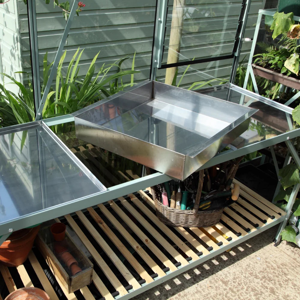 Metal table holds a reflective tray in a Rhino Greenhouse. Below, a wicker basket contains gardening tools. Surrounding plants and glass walls create a lush garden atmosphere.