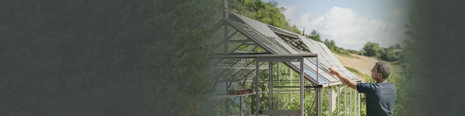 A person adjusts the roof of a glass Rhino Greenhouse on a sunny day, surrounded by lush greenery and hills. Pots with plants are visible inside.