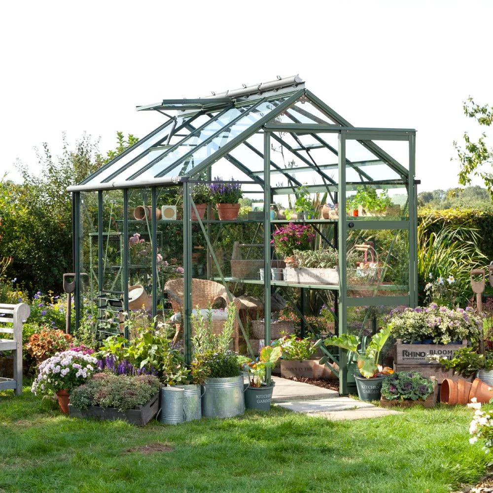 A green Rhino Greenhouse filled with potted plants and gardening tools stands amidst a lush garden. Metal containers labeled KITCHEN GARDEN and RHINO BLOOM are nearby.