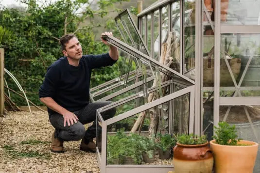 A person adjusts a window on a metal Rhino Greenhouse. Inside, plants grow; outside, potted herbs sit on gravel. The area is lush with greenery and natural light.