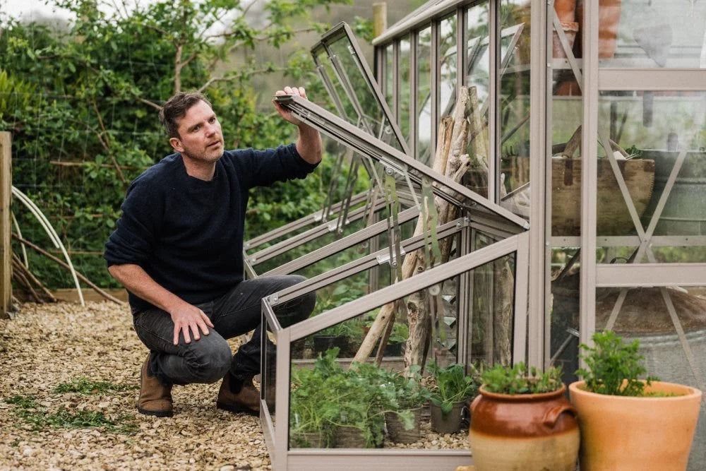 A man crouches, opening a vented window on a Rhino Greenhouse. Inside, plants are visible, while the exterior features gravel and clay pots with greenery, surrounded by lush vegetation.