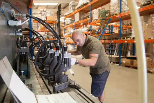 Worker operates industrial machinery in a warehouse with organized shelves filled with boxes and supplies. The scene suggests manufacturing or assembly within a well-stocked storage facility.