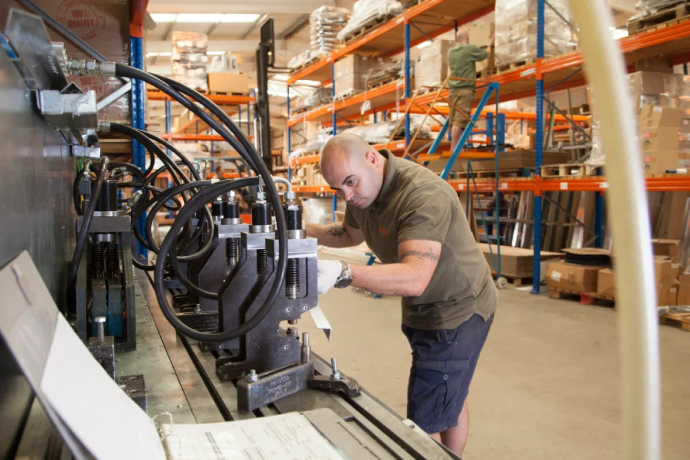 A person operates a machine with hoses in a warehouse filled with organized shelves containing boxes and tools. Another worker stands on a ladder in the background.