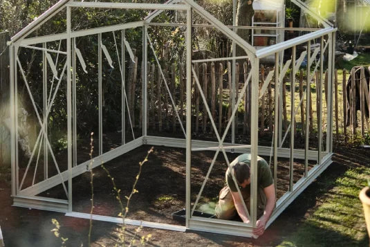 Man assembling Rhino Greenhouse frame, kneeling and adjusting a section in a sunny backyard with wooden fence and greenery.