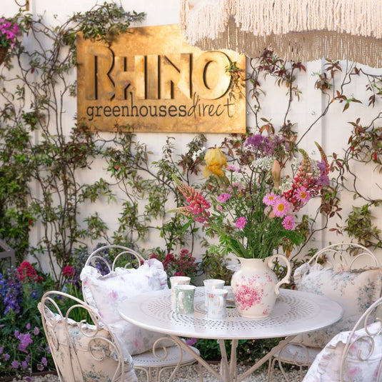 A white metal table hosts a floral pitcher and cups, surrounded by vintage chairs with cushions. The backdrop features climbing plants and a sign reading Rhino Rhino Greenhouses direct.