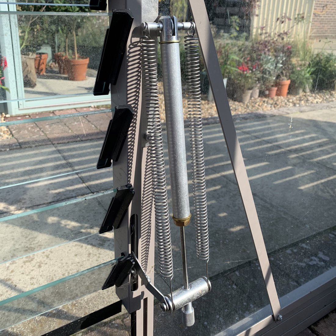 A metal Rhino Greenhouse window opener with springs is partially open, revealing potted plants and paving stones outside. Sunlight creates shadows on the transparent panels.