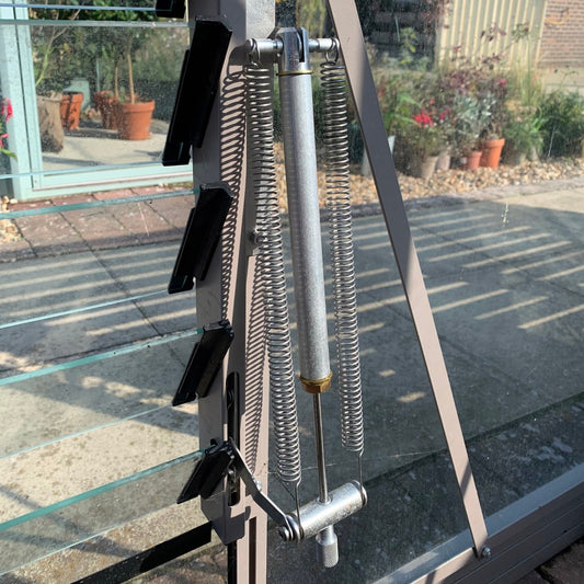 A metal Rhino Greenhouse window opener with springs is partially open, revealing potted plants and paving stones outside. Sunlight creates shadows on the transparent panels.