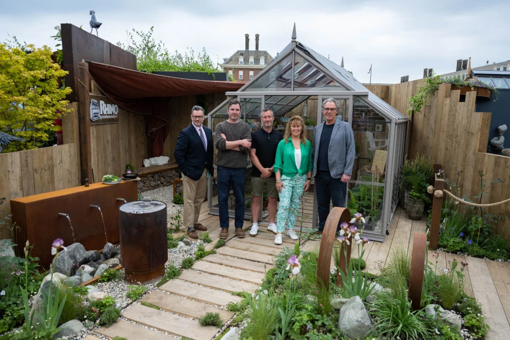A group of five people stands in front of a modern Rhino Greenhouse labeled Rhino, in a beautifully landscaped garden with wooden pathways and decorative plants.