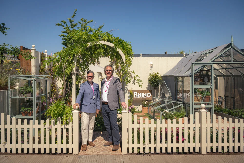 Two people stand smiling under an archway in a neatly maintained garden with Rhino Greenhouses. Behind them, a sign reads Rhino Rhino Greenhouses. The scene is enclosed by a white picket fence.
