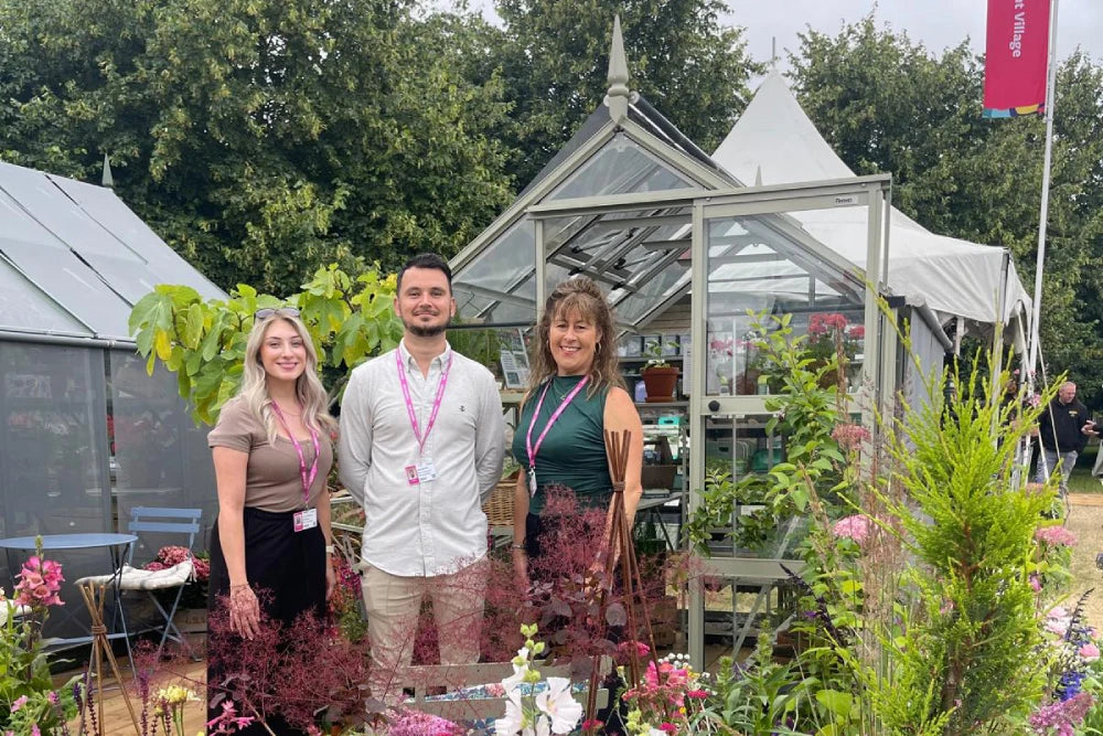 Rhino Greenhouse with three people standing in front, smiling, surrounded by vibrant plants and flowers. The setting is outdoors, with additional tents and greenery in the background.