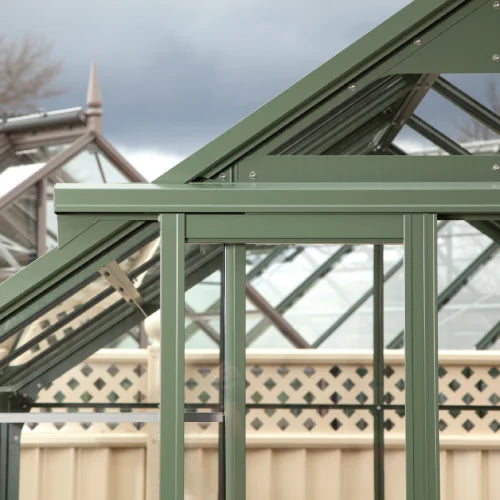 Rhino Greenhouse structure stands with green metal framing, showcasing glass paneling; neighboring Rhino Rhino Greenhouses and a beige fence visible in the background under a cloudy sky.