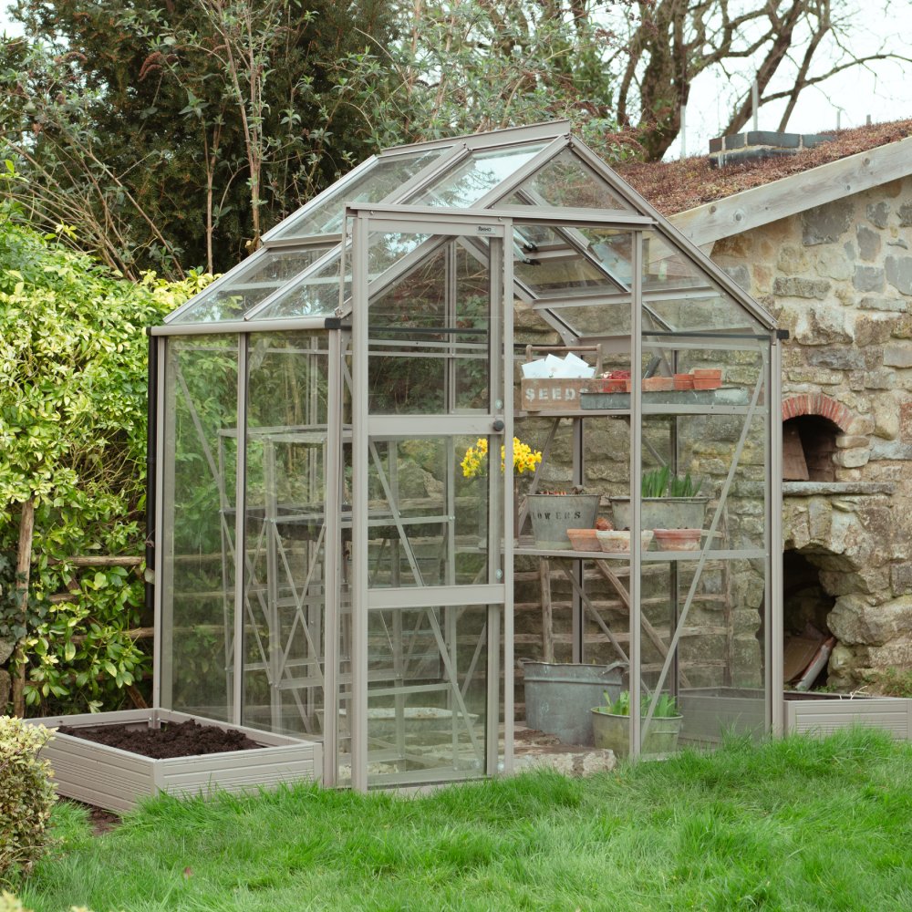 Rhino Greenhouse with metal frame and glass panels stands in a garden. Inside, shelves hold potted plants and gardening tools. Nearby, a stone building and lush greenery surround the scene.