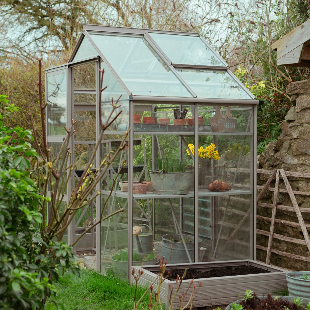 A glass Rhino Greenhouse holds potted plants and gardening tools, such as buckets and a watering can, surrounded by lush greenery and stone walls in a garden setting.