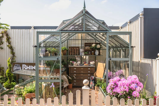 A Rhino Greenhouse stands open, displaying plants and gardening tools inside. Surrounded by a white fence and flowers, a Rhino Rhino Greenhouses sign is visible on a sunny day.