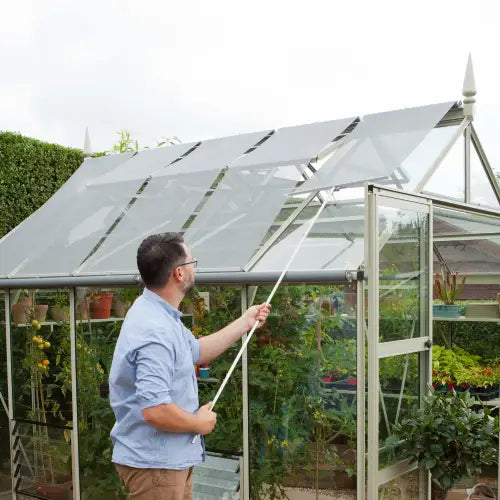 A person adjusts a vent with a pole on a glass Rhino Greenhouse. Inside, various plants are visible. The Rhino Greenhouse is situated in a garden, surrounded by greenery and hedges.