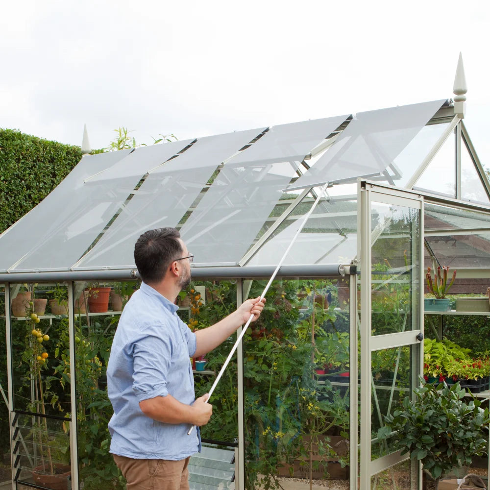 Man adjusting retractable shade on a glass Rhino Greenhouse using a pole; interior filled with thriving plants and gardening supplies, set in a lush garden environment.