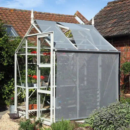 A white-framed Rhino Greenhouse with open vents houses plants inside. It stands on a gravel path beside a brick building, surrounded by greenery and flowerpots, under a clear sky.