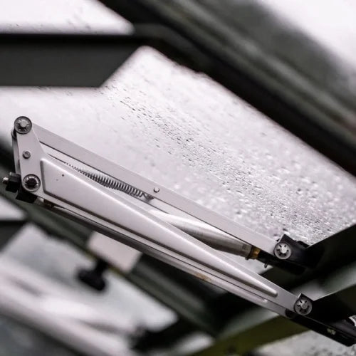 A metal hinge mechanism partially opens a Rhino Greenhouse window, surrounded by a rain-speckled surface.