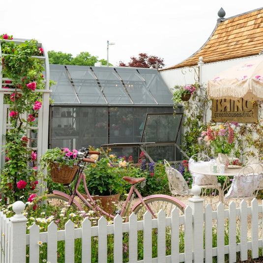 A pink bicycle with a flower-filled basket leans against a white picket fence. Behind it, a Rhino Greenhouse is surrounded by blooming plants and a quaint patio set next to a sign reading Rhino Rhino Greenhouses direct.