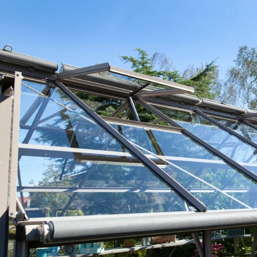 A Rhino Greenhouse features angled glass panels on a sturdy metal frame. It is set outdoors with trees in the background and a clear blue sky above, creating a serene environment.