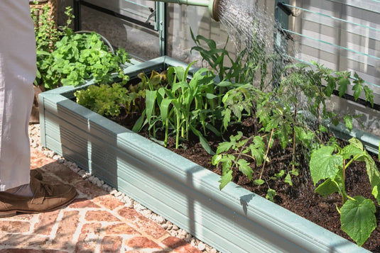 A person waters a variety of plants in a green Rhino metal raised garden bed, adjacent to a Rhino Greenhouse. The surrounding area features a brick and gravel pathway.