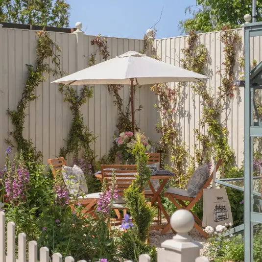 Wooden garden furniture with cushions sits under an umbrella among flowering plants, next to a Rhino Greenhouse. Tall plants climb a white fence, and a tote bag nearby reads Rhino.