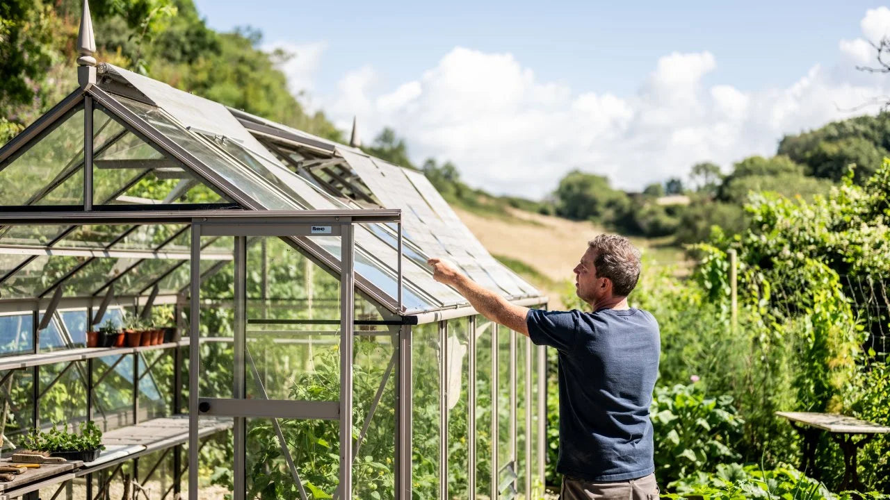 A man adjusts a glass pane on a Rhino Rhino Greenhouse filled with plants, set in a lush, hilly garden landscape under a bright blue sky.