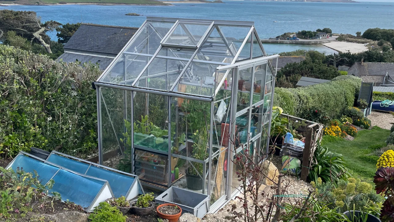 A glass Rhino Greenhouse shelters plants, surrounded by lush gardens and coastal scenery with a view of the ocean and neighboring buildings under a clear sky.