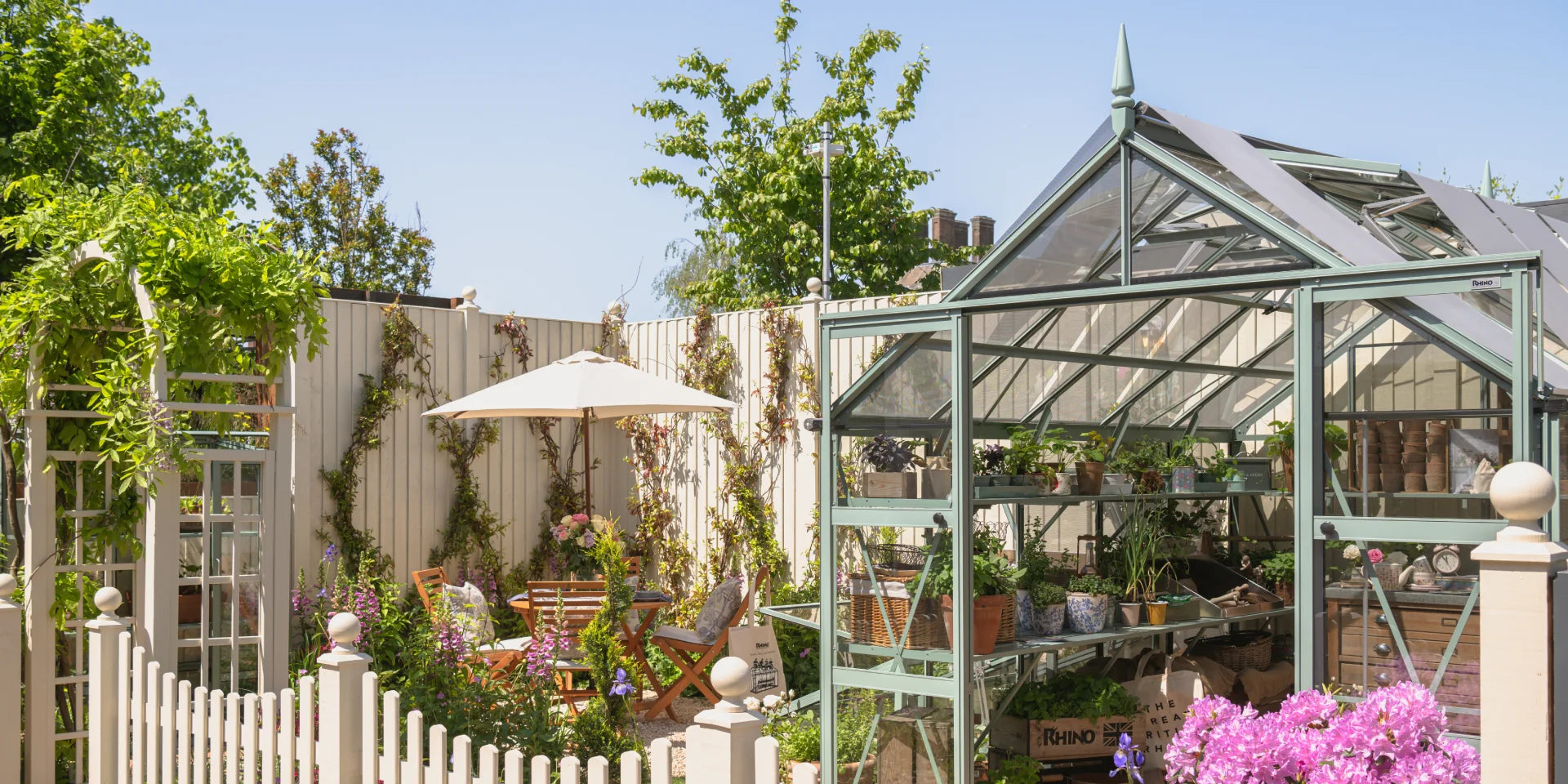 A glass Rhino Greenhouse filled with plants and gardening tools stands in a sunny garden, surrounded by flowers, a wooden fence, and outdoor furniture beneath a white umbrella. Rhino is displayed on the Rhino Greenhouse.