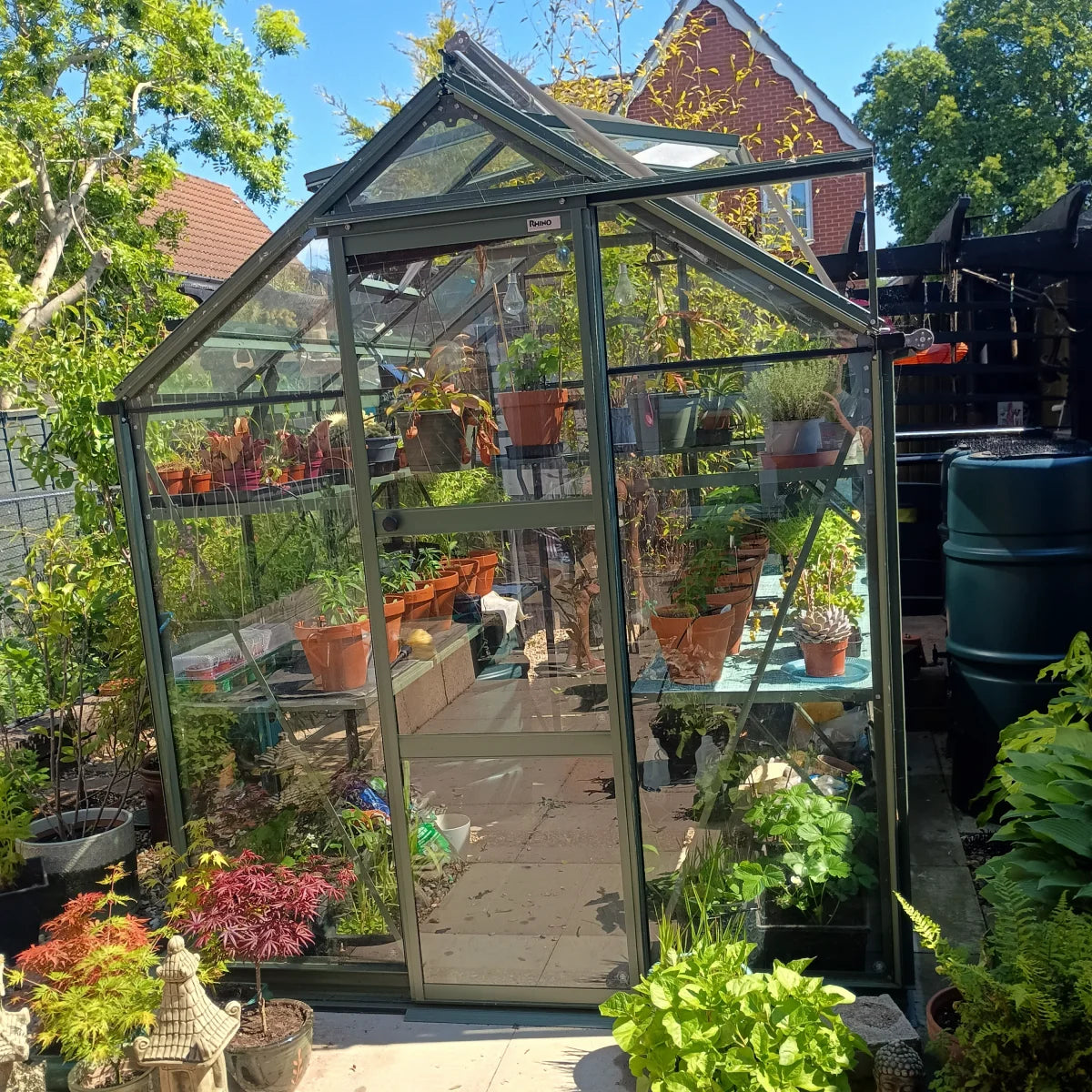 A glass Rhino Greenhouse filled with potted plants is situated in a garden, surrounded by lush greenery and other gardening items. It has a Rhino label on the top frame.