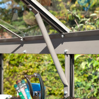 Metal support beam secures Rhino Greenhouse window, framed by bolts, in a lush garden setting. Gardening tools sit in the foreground, adding to the Rhino Greenhouse environment.