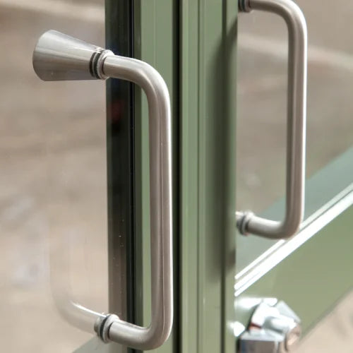 Metal door handles, affixed to a green-framed glass door, reflect light. The context suggests part of a Rhino Greenhouse structure, emphasizing durability and style in a gardening environment.