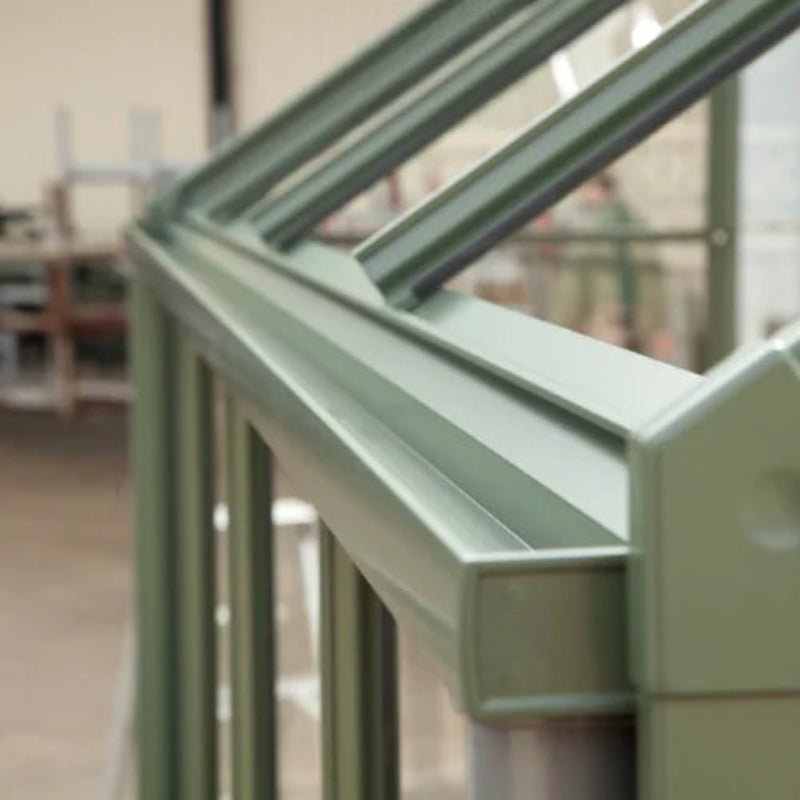 A green aluminum Rhino Greenhouse frame sits partially constructed indoors, with clear panels. The background shows a blurred workspace, suggesting assembly or manufacturing activities.