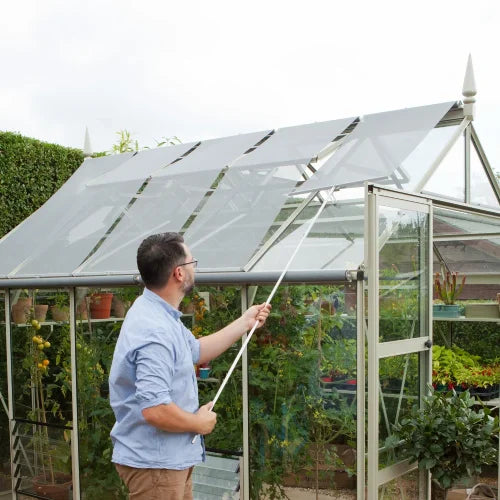 A man adjusts the shade of a glass Rhino Greenhouse with a rod. The Rhino Greenhouse, surrounded by plants, features open doors and a lush garden environment.