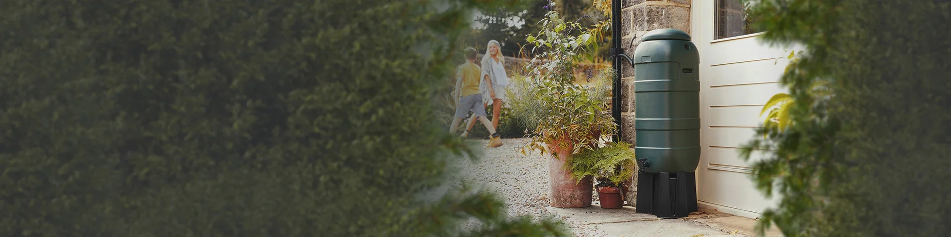 A green water tank stands beside potted plants near a building entrance, surrounded by greenery. Two people walk in the background on a gravel path.