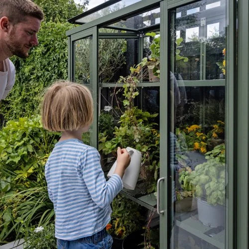 A child waters plants inside a small Rhino Greenhouse using a white watering can, observed by an adult. The Rhino Greenhouse is surrounded by lush greenery and colorful flowers.