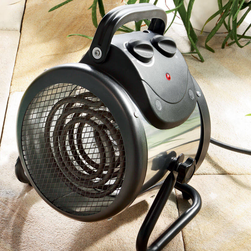 A portable electric heater with a cylindrical shape and front mesh grille sits on a tiled floor. It has a handle, two control knobs, and a red indicator light. Surrounding it are green plants.