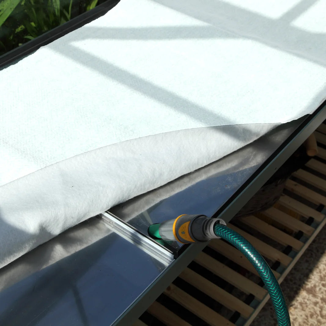 A garden hose nozzle is watering a table covered with a white cloth, positioned in a sunlit outdoor area on a wooden slatted surface.
